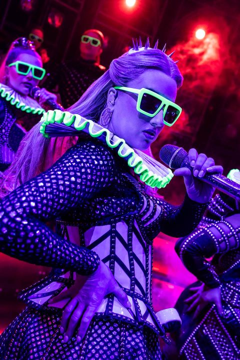 Performer in futuristic outfit and neon glasses sings into a microphone on a vibrant stage.