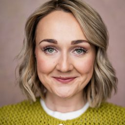 Smiling woman with short blonde hair, wearing a green textured top, against a soft pink background.